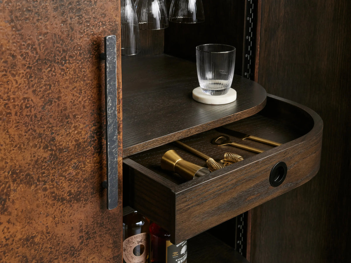 Ruiz Bar Cabinet: curved, dark oak and iron cabinet, one hammered copper door ajar. Interior displays hanging stemware, bottles, and wine rack.