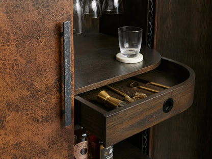 Ruiz Bar Cabinet: curved, dark oak and iron cabinet, one hammered copper door ajar. Interior displays hanging stemware, bottles, and wine rack.