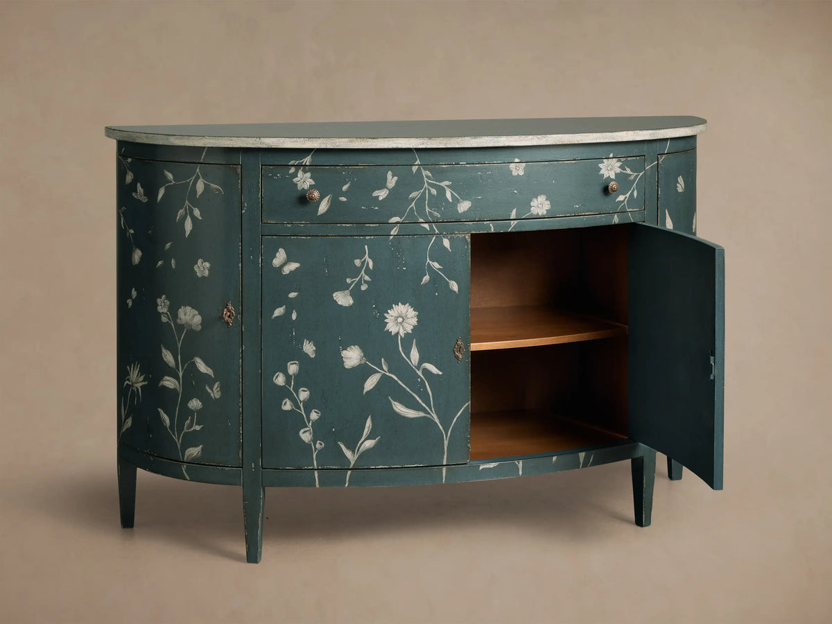 Distressed dark teal Sofia Bell'Arte Demilune Cabinet with white floral and butterfly motifs, light gray top, and open door to shelves.