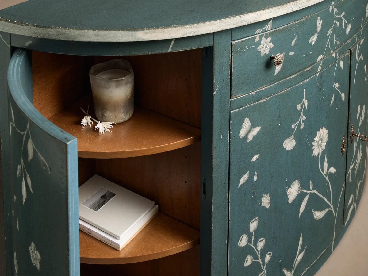 Close-up of the distressed teal Sofia Bell'Arte Demilune Cabinet with white floral motifs, one door open revealing wooden shelves.
