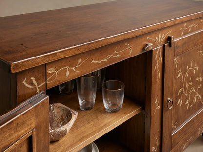Stefania Buffet: Warm brown wooden buffet with hand-painted gold foliage, left door ajar revealing two interior shelves.