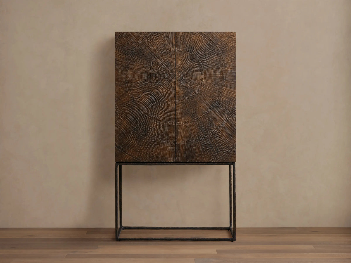 Theota Bar Cabinet with a dark brown sunburst pattern on teak, resting on slender black metal legs against a beige wall.