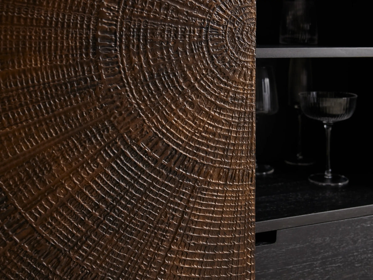 Theota Bar Cabinet with a dark brown sunburst pattern on teak, resting on slender black metal legs against a beige wall.