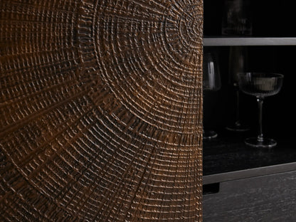 Theota Bar Cabinet with a dark brown sunburst pattern on teak, resting on slender black metal legs against a beige wall.