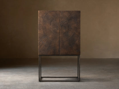 Theota Bar Cabinet with a dark brown sunburst pattern on teak, resting on slender black metal legs against a beige wall.