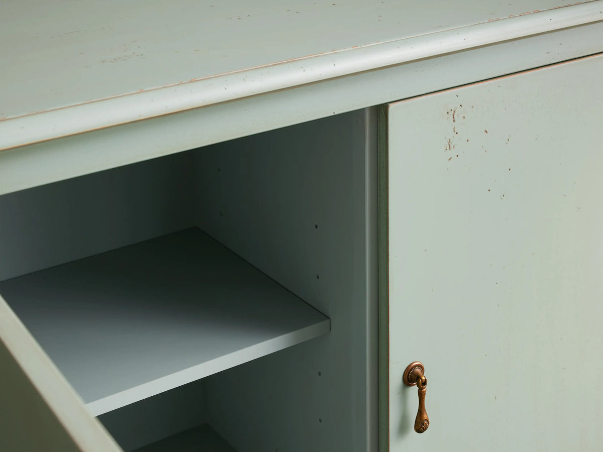 Varzi Cabinet in light green with two doors, featuring antique pendant pull handles. Curved legs and distressed finish on beige background.