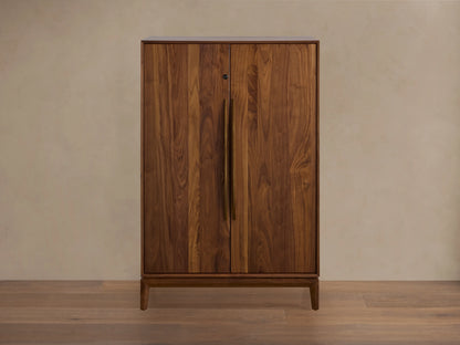 Willard Bar Cabinet, a rich dark brown walnut wood cabinet with two doors and long vertical brass handles, standing on a wooden floor.