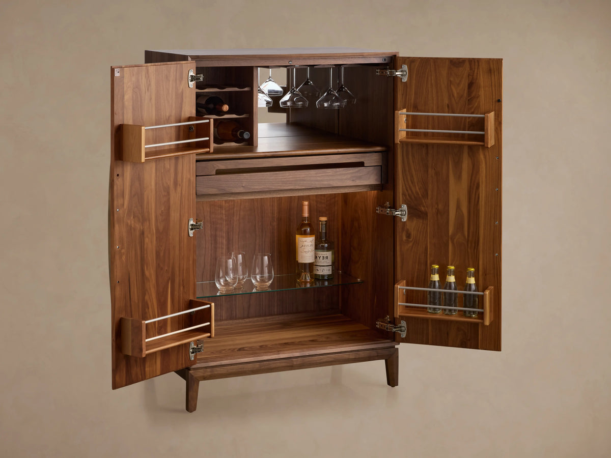 Willard Bar Cabinet, a rich dark brown walnut wood cabinet with two doors and long vertical brass handles, standing on a wooden floor.