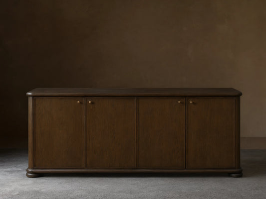 Warby Sideboard made from solid oak wood with a rich mocha finish. Features four cabinet doors with brass hardware on a smooth wood floor.