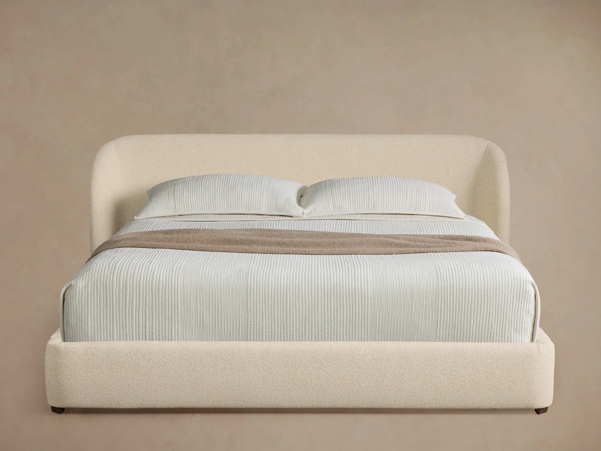 Adalynn Bed: Cream boucle upholstered bed with curved headboard, light striped bedding, and a taupe throw on wood floor.