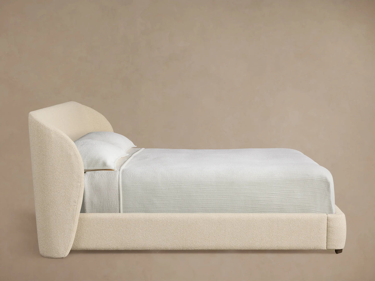 Close-up of the Adalynn Bed's upholstered headboard, showing a rounded corner in light beige textured fabric.