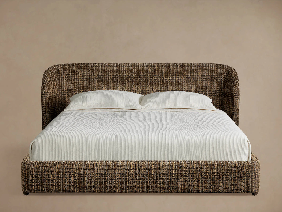 Adalynn Bed with a brown & black tweed-like upholstered, curved headboard & low frame, dressed with white bedding & pillows, on wood floor.