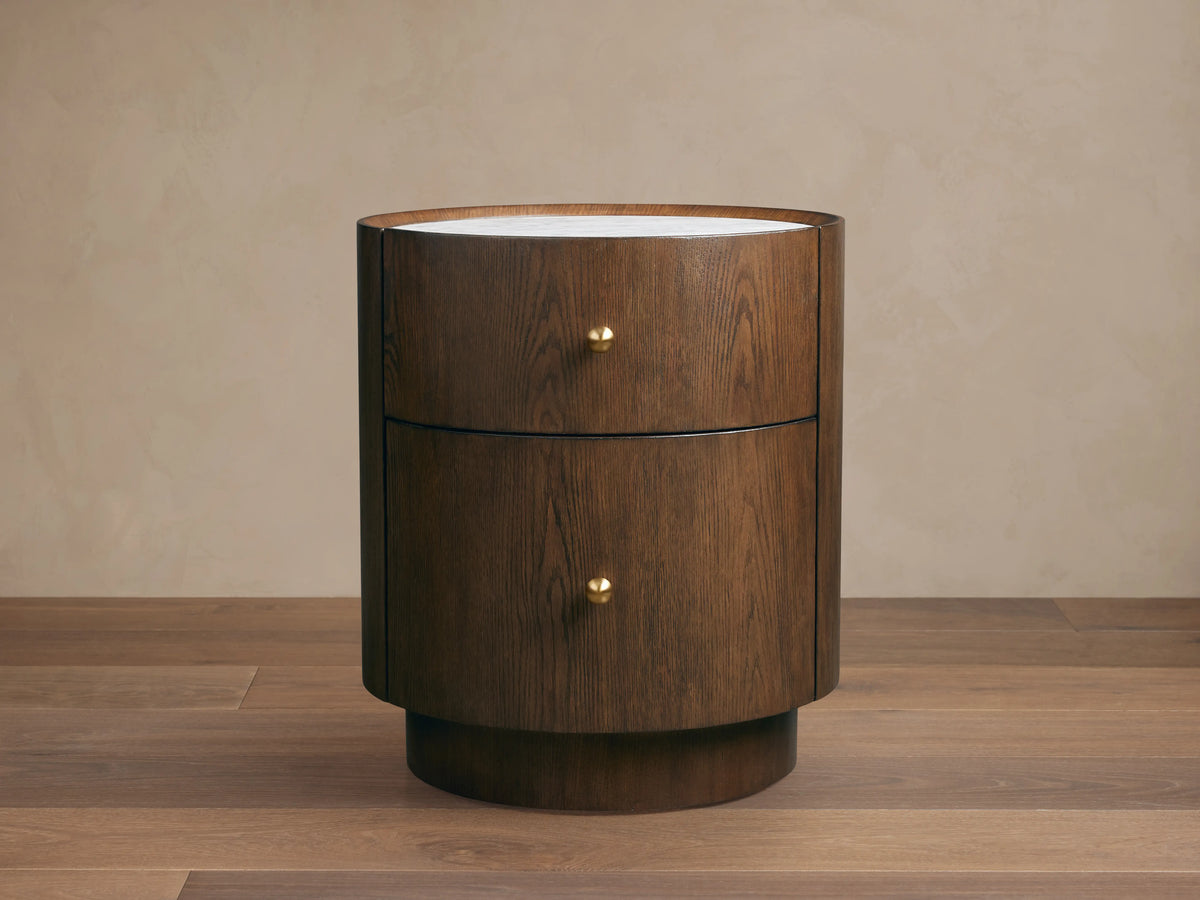 Round Ashbrook Nightstand with mocha oak veneer, two drawers, brass knobs, and white Carrara marble top on wood floor.