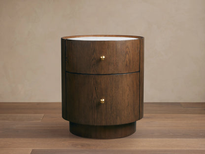 Round Ashbrook Nightstand with mocha oak veneer, two drawers, brass knobs, and white Carrara marble top on wood floor.