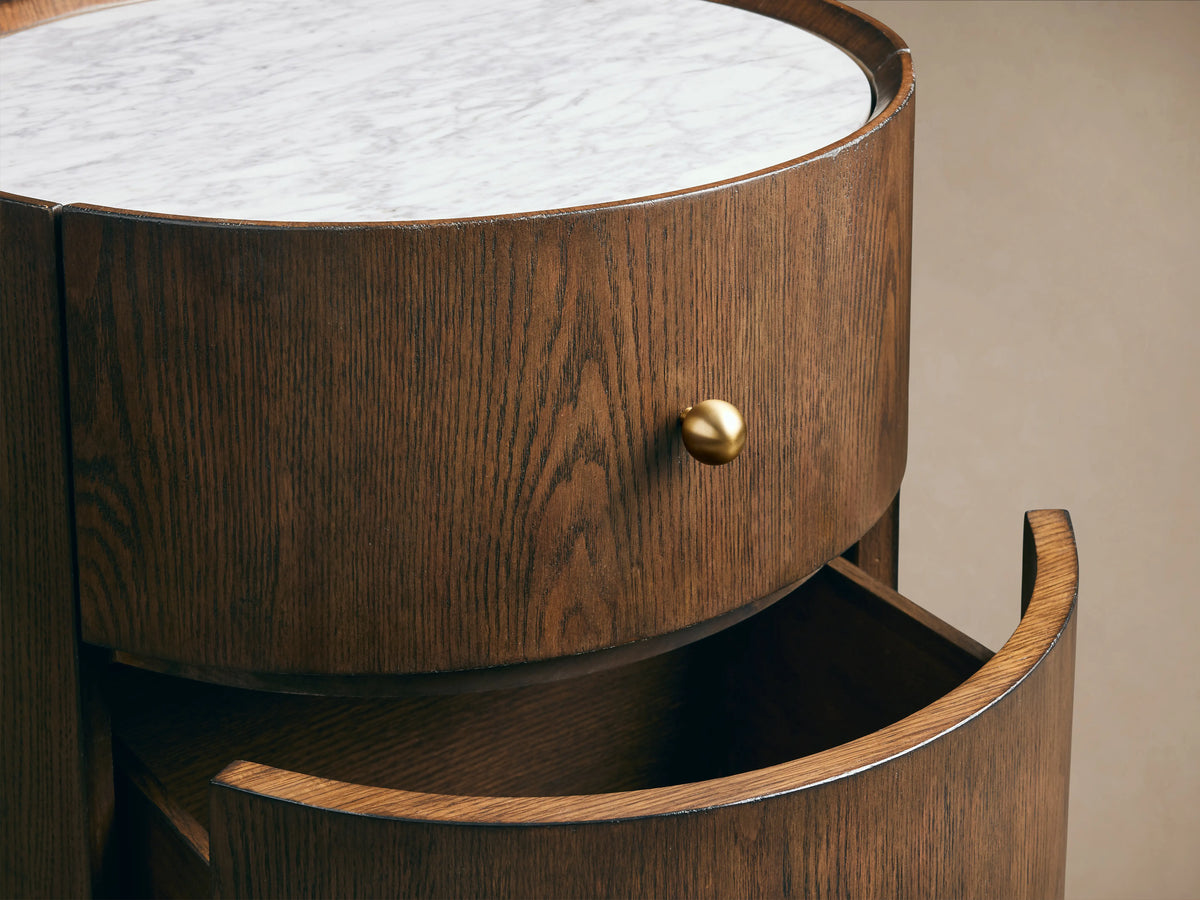 Round Ashbrook Nightstand with mocha oak veneer, two drawers, brass knobs, and white Carrara marble top on wood floor.