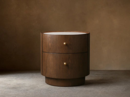 Round Ashbrook Nightstand with mocha oak veneer, two drawers, brass knobs, and white Carrara marble top on wood floor.