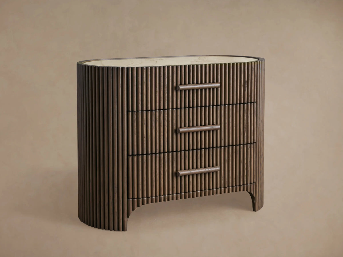 Astor Nightstand: Dark brown reeded oak oval body, light natural marble top, three drawers with horizontal wooden pulls, on wood floor.