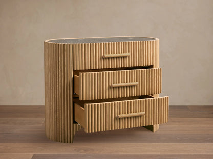 Astor Nightstand: Light oak with vertical reeding, 3 drawers, wooden pulls, and a dark marble top, on a wood floor.