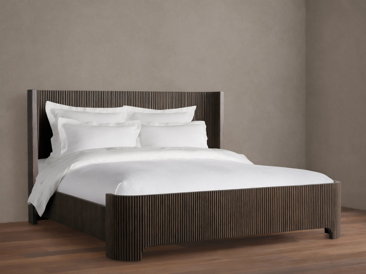 Astor Bed with solid oak reeding, dark finish. White bedding with pillows contrasts the vertical slats, set on wooden floor.