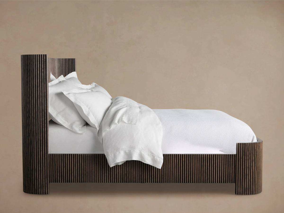 Astor Bed in side view showing oak veneer frame with solid reeding detail, adorned with white bedding and pillows, against a beige background.