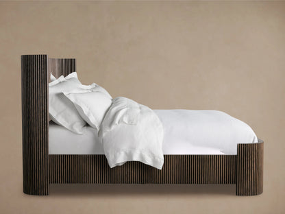 Astor Bed in side view showing oak veneer frame with solid reeding detail, adorned with white bedding and pillows, against a beige background.
