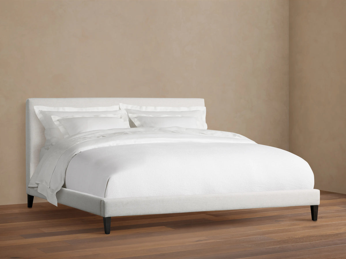 Beck Bed with a white fabric-covered frame, simple headboard, and dark wooden legs, set against a light wall and wood floor.