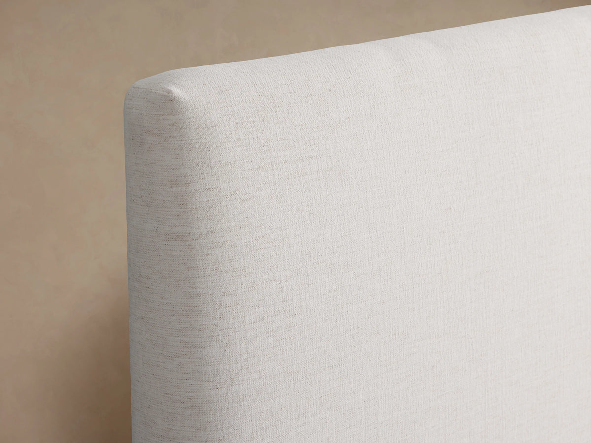 Beck Bed with a white fabric-covered frame, simple headboard, and dark wooden legs, set against a light wall and wood floor.