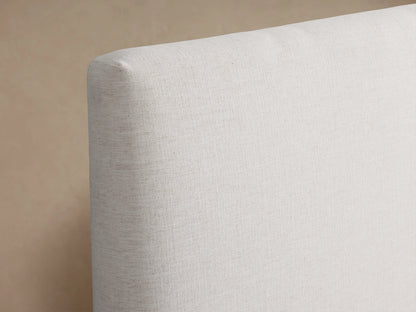 Beck Bed with a white fabric-covered frame, simple headboard, and dark wooden legs, set against a light wall and wood floor.