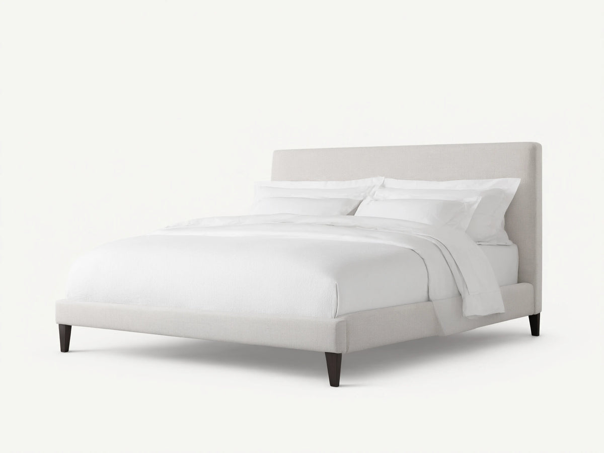 Beck Bed with a white fabric-covered frame, simple headboard, and dark wooden legs, set against a light wall and wood floor.