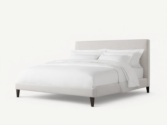 Beck Bed with a white fabric-covered frame, simple headboard, and dark wooden legs, set against a light wall and wood floor.