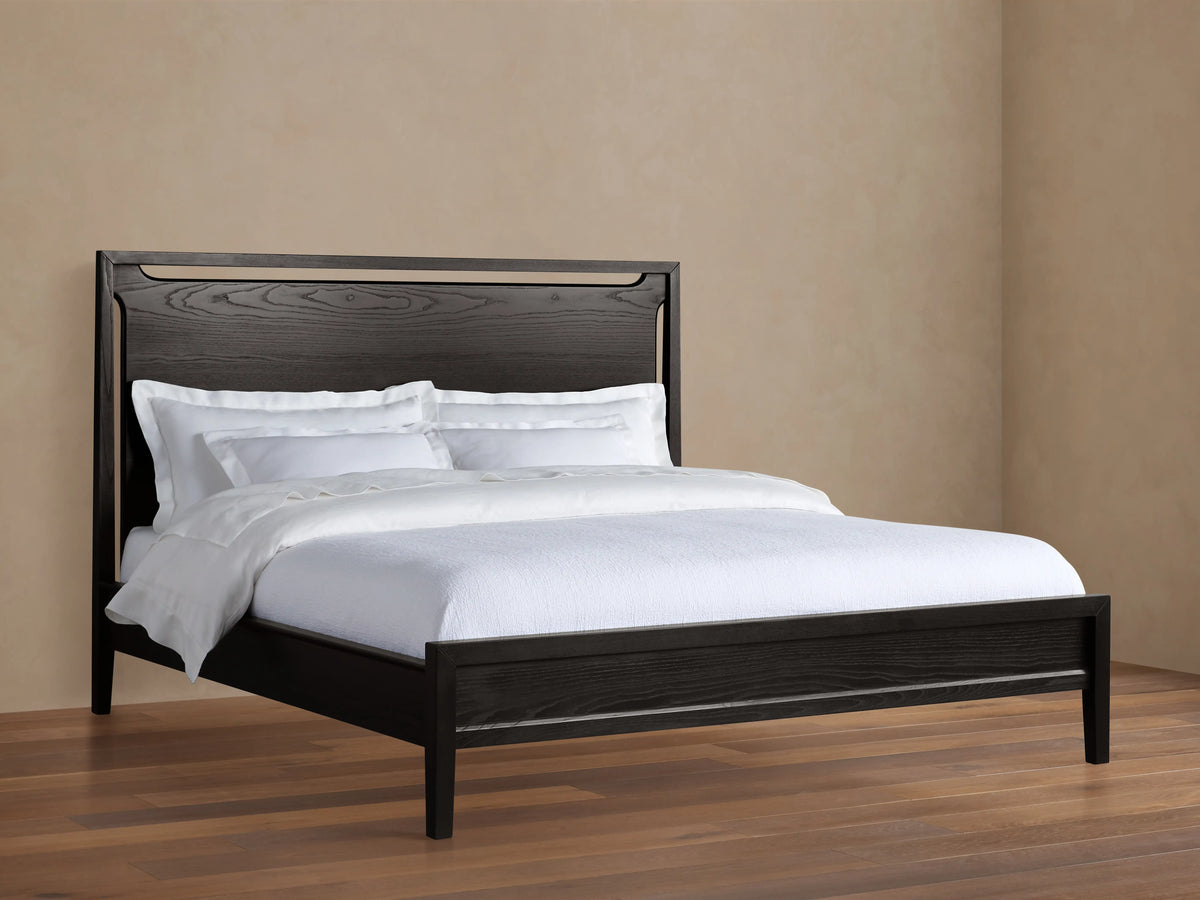 Ebony-colored Brock Bed with a high solid ash wood headboard and white bedding, set on a wooden floor in a beige room.