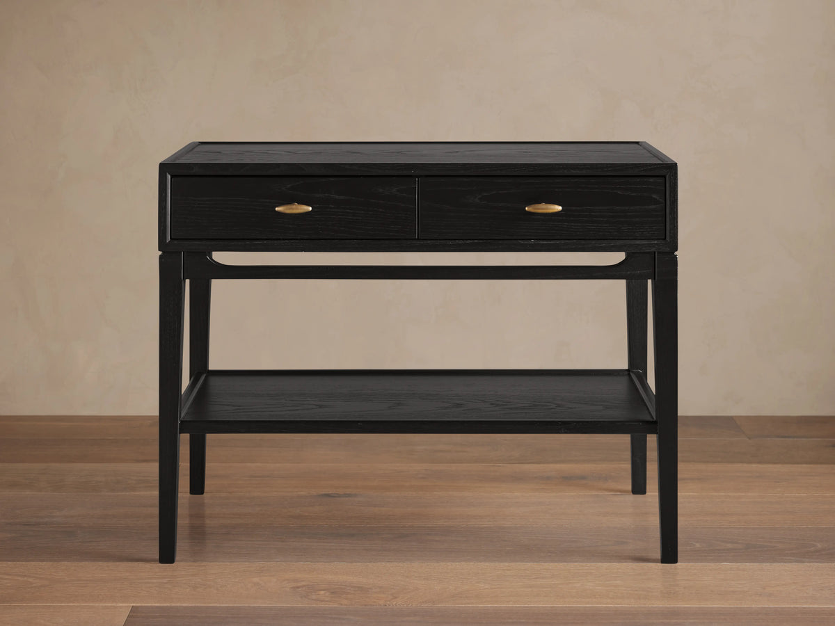 Brock Nightstand in ebony ash wood with two brass-handled drawers and lower shelf, set on flat wood flooring against beige wall.