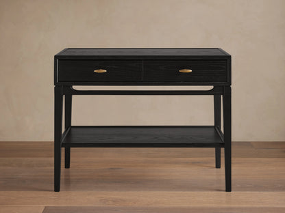 Brock Nightstand in ebony ash wood with two brass-handled drawers and lower shelf, set on flat wood flooring against beige wall.