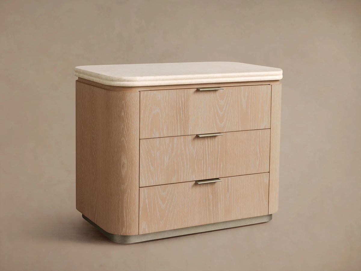 Caden Nightstand, sand finish oak veneer, light stone top, 3 drawers with pewter edge-pulls, on wood floor.