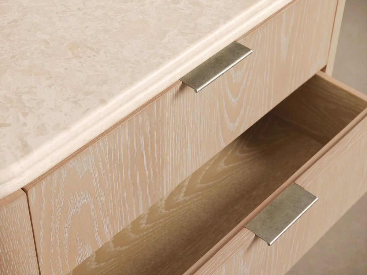 Caden Nightstand: sand oak veneer, cream stone top, three drawers with pewter edge-pulls, rounded sides, gray plinth.