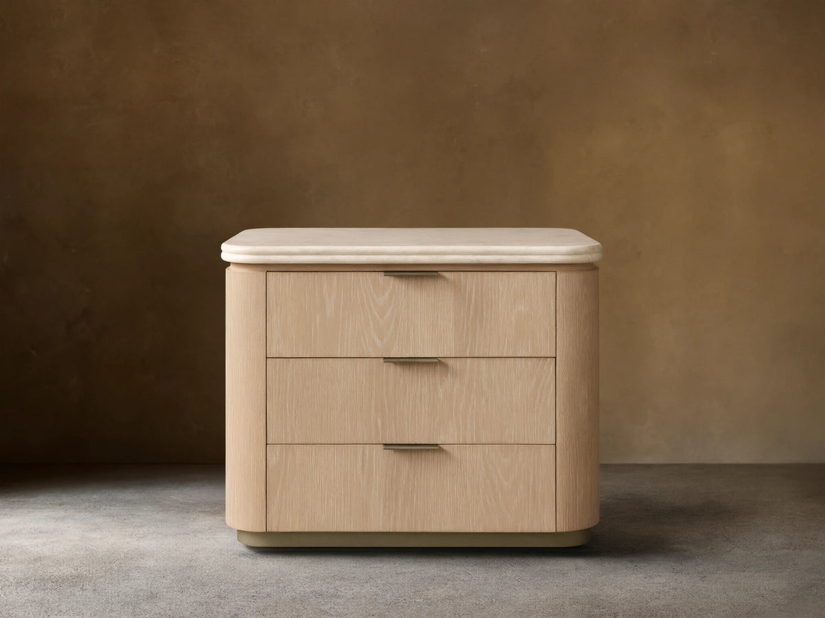 Caden Nightstand, sand finish oak veneer, light stone top, 3 drawers with pewter edge-pulls, on wood floor.