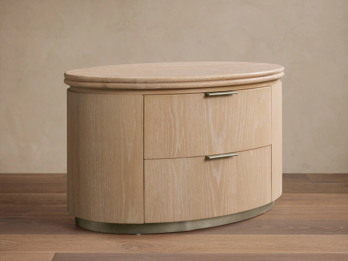 Caden Wood Oval Nightstand in sand oak veneer with an oval top, curved front, 2 drawers & pewter handles, on wood floor.