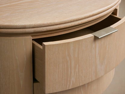 Caden Wood Oval Nightstand in sand finish oak veneer, shown with one curved drawer partially open, featuring pewter edge-pull hardware.