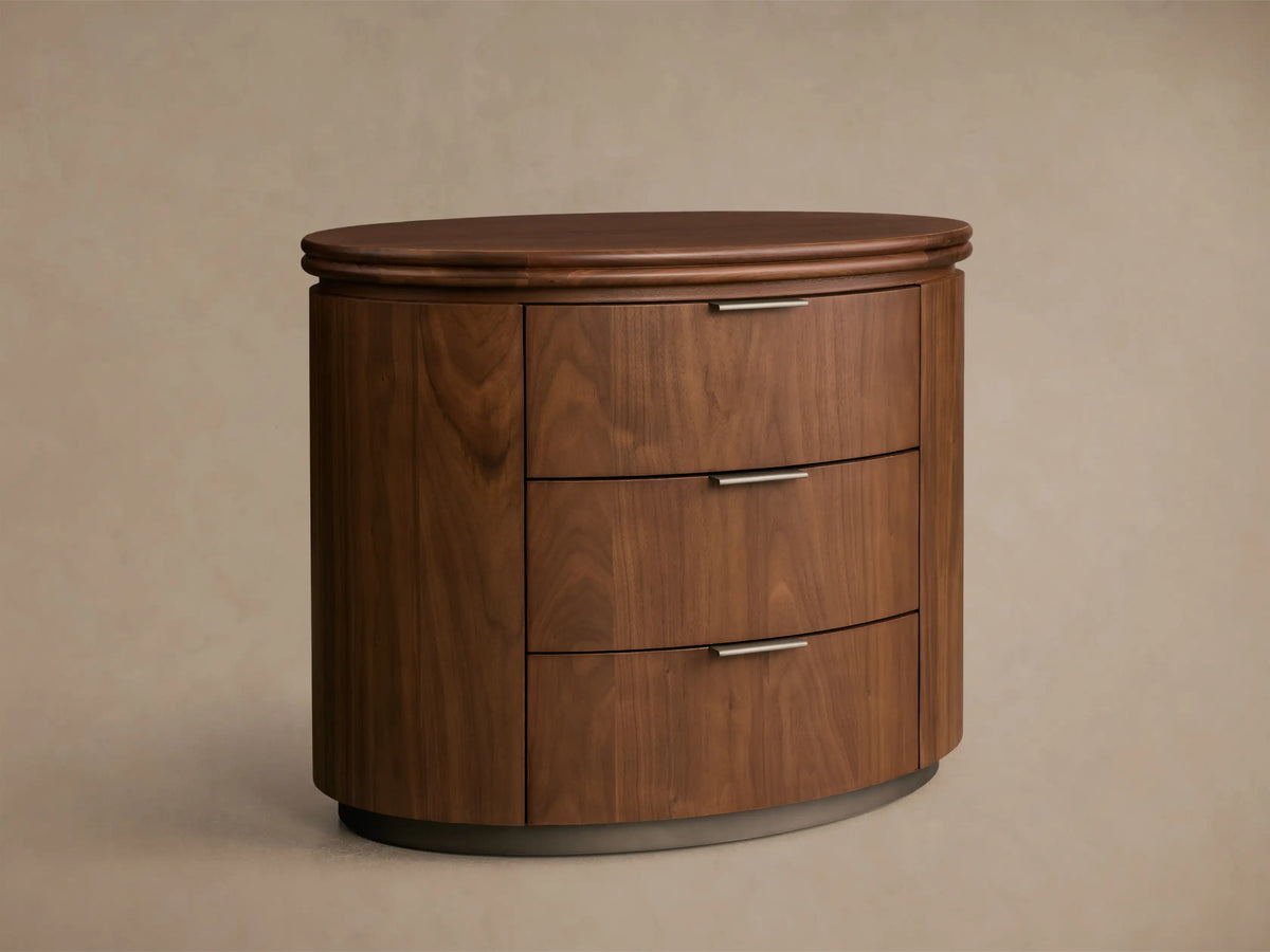 Front view of the walnut Caden Wood Oval Nightstand, an oval, dark wood piece with three drawers and antique bronze bar pull handles.