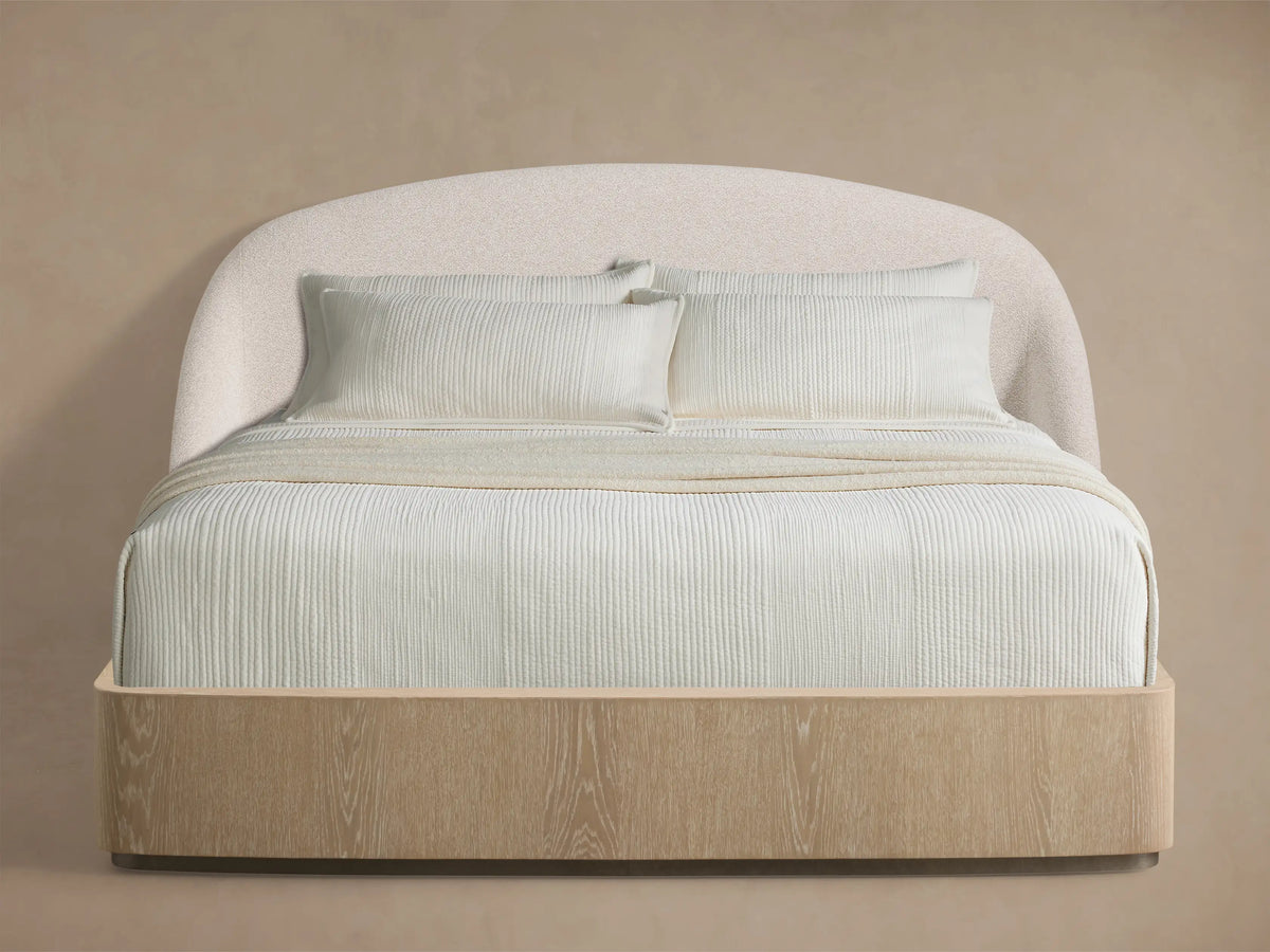 Front view of the Caden Bed with a textured cream upholstered curved headboard, light oak-finish wood frame, and white ribbed bedding.