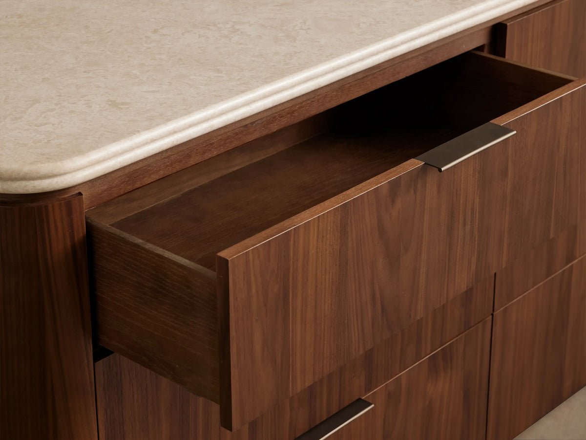 Caden Six-Drawer Dresser in light walnut wood with a white stone top, six drawers with antique bronze pulls, against a brown wall.