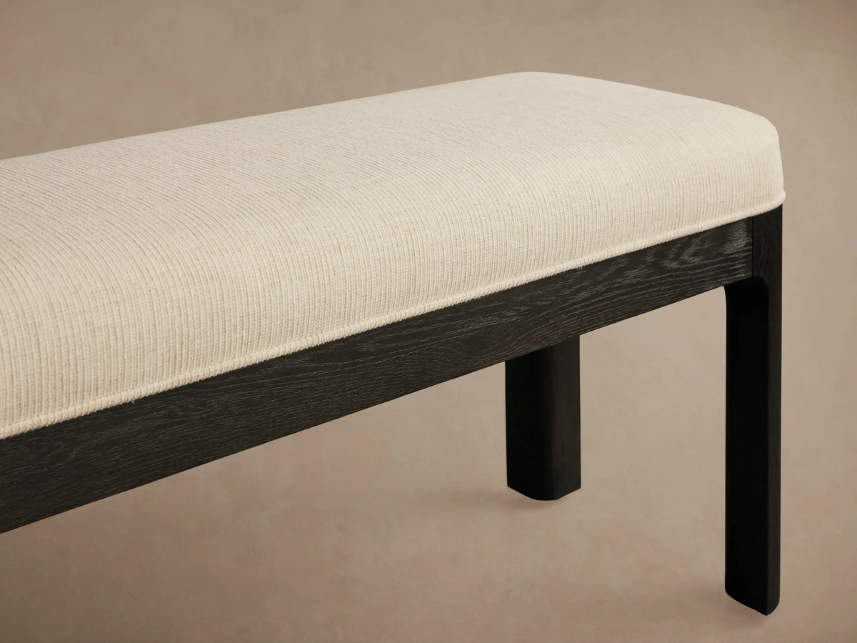 Calista Bench: Minimalist, long bench with rounded cream upholstered top and dark wood frame, on a wooden floor.