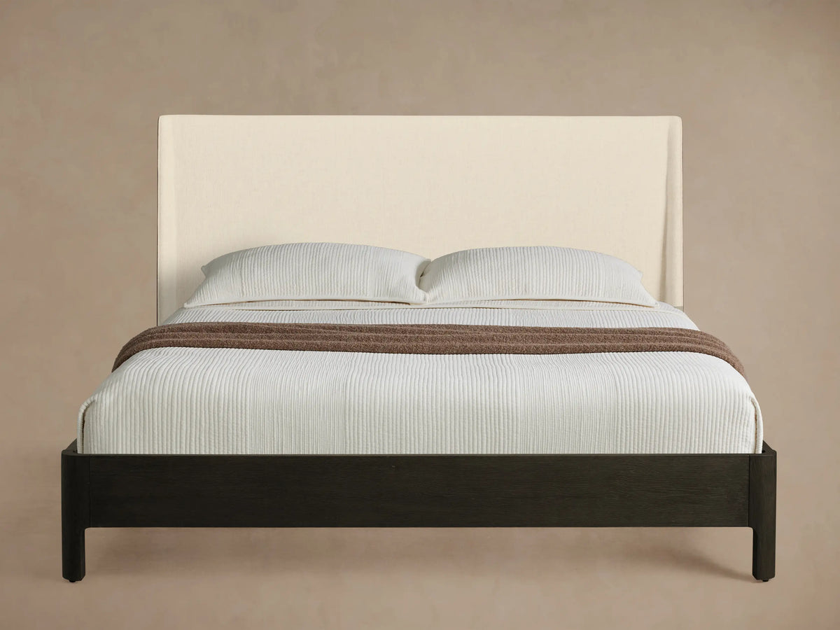 Calista Bed with cream upholstered headboard, dark brown wood frame, white textured bedding, and a brown throw.