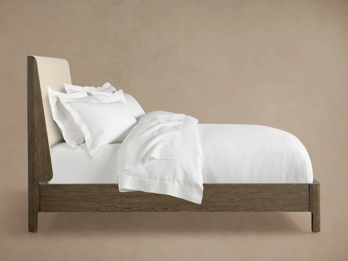 Close-up of the upholstered headboard of the Calista Bed, showing light beige textured fabric and dark wood frame edge.