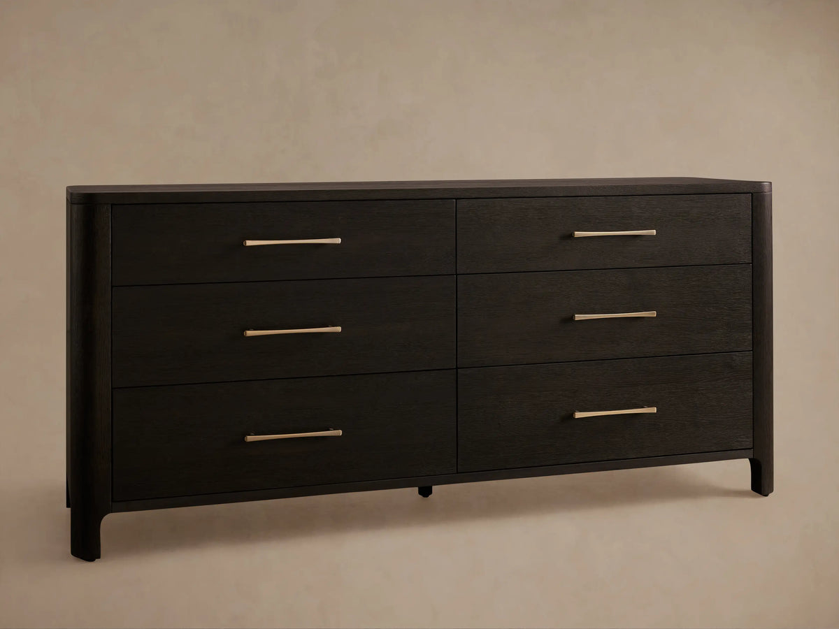 Calista Six-Drawer Dresser close-up. Dark oak with visible grain, rounded left corner. Lower drawer open. Bronze-gold bar handles.