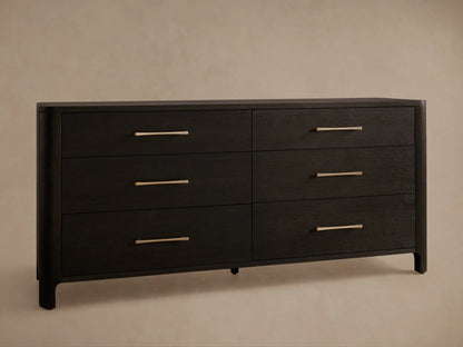 Calista Six-Drawer Dresser close-up. Dark oak with visible grain, rounded left corner. Lower drawer open. Bronze-gold bar handles.