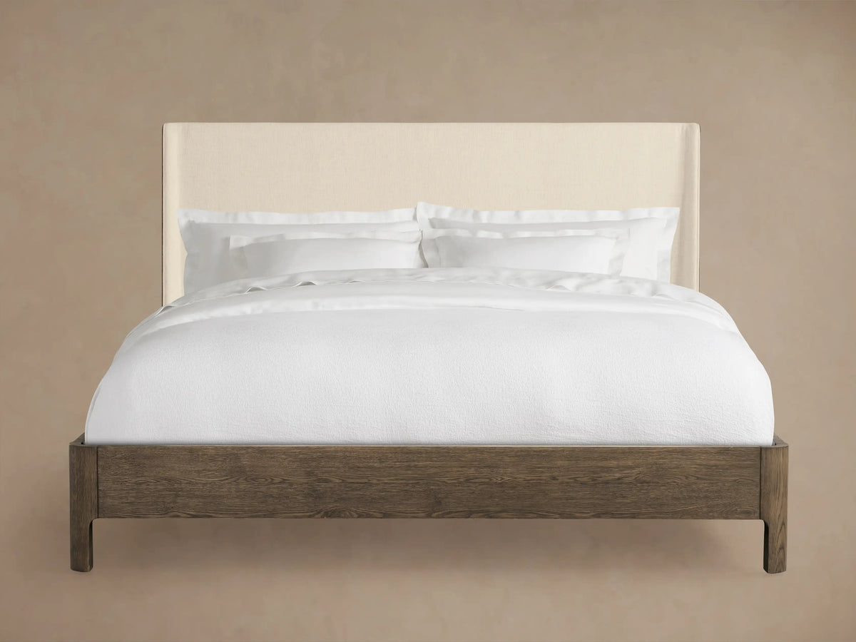 Calista Bed with upholstered beige headboard and dark oak frame, made up with white sheets and pillows, on a wooden floor.