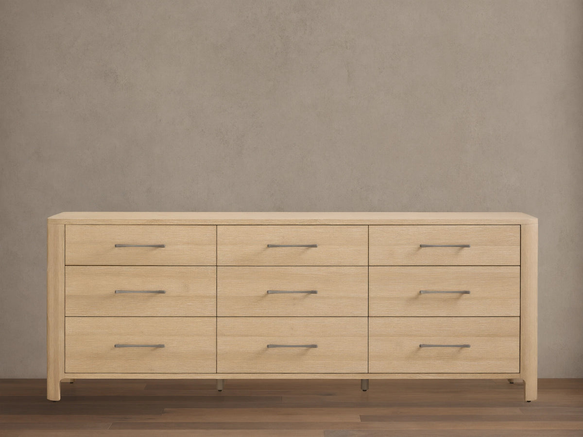 Calista Nine-Drawer Dresser: light oak wood with rounded edges. Features nine drawers with horizontal matte silver bar pulls. Front view.