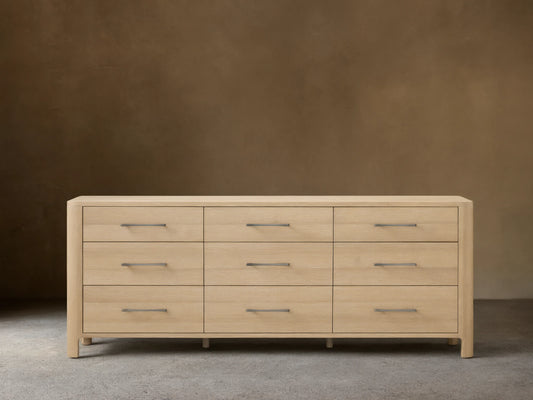 Calista Nine-Drawer Dresser: light oak wood with rounded edges. Features nine drawers with horizontal matte silver bar pulls. Front view.