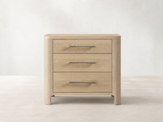 Calista Closed Nightstand in light oak wood with prominent grain, featuring rounded edges and three drawers with gray pull handles.